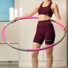 Na zdjęciu widoczna jest osoba trzymająca hula hop w kolorze szaro-różowym. Osoba ubrana jest w bordowy strój sportowy z napisem "GLAMOUR".