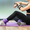 Osoba siedzi na fioletowej macie do ćwiczeń, nosząc obciążniki na rękach i kostkach z widocznym napisem "DRFIT 1 KG". Na stopach ma białe buty sportowe z czarnymi i szarymi wzorami.
