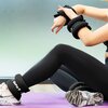 Na zdjęciu widać osobę siedzącą na fioletowej macie do ćwiczeń, z obciążnikami na rękach i kostkach, na których widnieje napis "DRFIT". Osoba ma na sobie czarne legginsy i białe buty sportowe z czarnymi i szarymi wzorami.