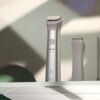 Na zdjęciu widoczny jest srebrny trymer z napisem "PHILIPS All-in-One" oraz "Wet & Dry", stojący na szklanej półce. W tle znajduje się drugi trymer, częściowo widoczny.