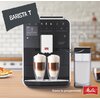 Ekspres do kawy z wyświetlaczem i przyciskami dotykowymi, na którym widnieje napis "Please select your beverage", przygotowuje dwie kawy z mleczną pianką w szklankach. Obok ekspresu znajduje się pojemnik na mleko, a na zdjęciu widoczne są napisy "BARISTA 