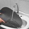 Na zdjęciu widać szarą podkładkę na stół, która jest myta pod bieżącą wodą w zlewie. Woda spływa po powierzchni podkładki, a w tle widoczna jest bateria zlewozmywakowa.