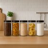 Na zdjęciu znajduje się pięć szklanych pojemników z czarnymi pokrywkami, wypełnionych różnymi produktami spożywczymi: ziarnami kawy, kolorowym makaronem, płatkami owsianymi, makaronem w kształcie gniazd i białym proszkiem. W tle widoczna jest biała ściana