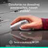 Na zdjęciu widać dłoń trzymającą ciemnoszarą mysz komputerową na gładkiej powierzchni, obok laptopa. Na zdjęciu znajduje się tekst: "Działanie na dowolnej powierzchni, nawet na szkle" oraz "Teraz z czułością 8K DPI".