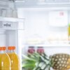 Wnętrze lodówki z widocznymi półkami, na których znajdują się butelki z pomarańczowym napojem, słoiki oraz ananas. Na drzwiach lodówki widoczny jest napis "vestfrost".