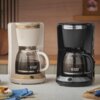 Na zdjęciu znajdują się dwa ekspresy do kawy marki Russell Hobbs, jeden w kolorze beżowym, a drugi czarnym, z przezroczystymi dzbankami wypełnionymi kawą. Na dzbankach widoczne są miarki, a na ekspresach logo "Russell Hobbs".