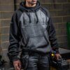 Mężczyzna ubrany w szaro-czarną bluzę z kapturem, na której widnieje napis "DEWALT GUARANTEED TOUGH", trzyma w ręku czarną skrzynkę narzędziową. Na rękawie bluzy znajduje się napis "GUARANTEED TOUGH".