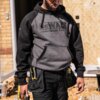 Mężczyzna ubrany w szaro-czarną bluzę z kapturem, na której widnieje napis "DEWALT GUARANTEED TOUGH", stoi na tle budynku z cegły. Na jego pasie znajdują się narzędzia w czarnych kieszeniach.