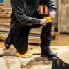 Osoba klęczy na drewnianej powierzchni, używając żółtych nakolanników roboczych DEWALT DWC15-001, ubrana w ciemne spodnie i bluzę z widocznym logo DEWALT. W tle znajdują się drewniane deski i kałuża wody.