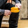 Osoba wkłada żółty nakolannik do kieszeni na kolanie czarnych spodni roboczych. Na nakolanniku widoczne są napisy: "DEWALT" oraz "GUARANTEED TOUGH".