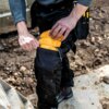Osoba wkłada żółty ochraniacz kolan do kieszeni na spodniach roboczych. Na ochraniaczu widoczny jest napis "DEWALT".