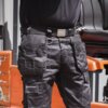 Czarna odzież robocza z wieloma kieszeniami, w tym pasek z metalową klamrą z logo "DEWALT". W tle widoczny jest fragment pomarańczowego sprzętu przemysłowego.