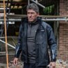 Na zdjęciu widoczny jest mężczyzna w czarnej kurtce roboczej z logo "DEWALT" na piersi, stojący na tle rusztowania i ceglanej ściany. Kurtka ma odblaskowe elementy na rękawach.