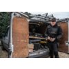 Mężczyzna w kurtce roboczej DEWALT Softshell Tough DWC263-001-M stoi obok otwartego bagażnika samochodu dostawczego, w którym znajdują się narzędzia i materiały budowlane. Na kartonie widoczny jest napis "FRAGILE".