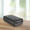 Czarny powerbank leży na drewnianej powierzchni, posiada dwa porty USB, jeden port USB-C oraz cztery diody LED wskazujące poziom naładowania. W tle widoczne jest rozmyte, jasne tło.