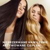 Dwie osoby z długimi, lśniącymi włosami, jedna z ciemnymi, druga z jasnymi, na tle beżowej ściany. Na dole zdjęcia znajduje się tekst: "NIEPRZERWANE NAWILŻENIE AKTYWOWANE CIEPŁEM* *wyłącznie w przypadku używania razem z kremem HEAT & GLOW".