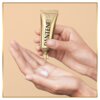 Złota tubka z napisem "PANTENE PRO-V" jest trzymana nad otwartą dłonią, na której znajduje się niewielka ilość białego kremu. Tło jest jednolite, w kolorze beżowym.