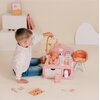 Na zdjęciu znajduje się zestaw zabawkowy "Kącik opiekunki SMOBY Baby Nurse 7600220393" w kolorach różowym i białym, z lalką leżącą na przewijaku oraz akcesoriami do opieki. W tle widoczne są białe i różowe elementy dekoracyjne oraz książeczki.