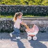 Na zdjęciu widoczny jest różowy wózek dziecięcy z napisem "BABY NURSE" na tle kamiennego muru i trawnika. Wózek ma białe koła i żółty uchwyt.