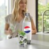 Na zdjęciu znajduje się butelka z napisem "Pepsi Cola" oraz szklanka z napojem gazowanym, w której znajdują się jagody i liście mięty. W tle widoczna jest osoba w jasnej bluzce, a za nią okno z widokiem na zieleń.