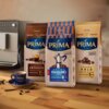 Na zdjęciu znajdują się trzy opakowania kawy PRIMA: Aroma Intenso, Aroma Italiano i Aroma Gold, ustawione na drewnianym blacie obok filiżanki z logo PRIMA. Na opakowaniach widoczne są informacje o intensywności kawy oraz jej rodzaju, a w tle znajduje się 