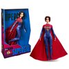 Na zdjęciu znajduje się lalka w stroju Supergirl z czerwonym płaszczem i niebieskim kostiumem, obok której widoczne jest opakowanie z napisem "Barbie Signature" oraz "SUPERGIRL". Opakowanie ma niebiesko-czerwone tło z grafiką przedstawiającą tę samą posta