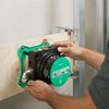 Na zdjęciu widoczny jest zielony korpus Boxa HANSGROHE iBox 01500180, trzymany przez osobę, zamontowany na drewnianej płycie z metalowymi elementami konstrukcyjnymi. Na zielonej pokrywie znajduje się logo "hansgrohe" oraz ilustracje montażowe.
