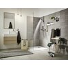 Nowoczesna łazienka z prysznicem wyposażonym w słuchawkę prysznicową HANSGROHE Pulsify Select S 24111000 Chrom, na ścianie znajdują się półki z kosmetykami i rośliną. Obok prysznica stoi krzesło z ręcznikami, a na podłodze leży dywanik i kosz na pranie.