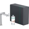 Na zdjęciu znajduje się chromowana bateria kuchenna, obok której stoi butelka z napisem "hansgrohe claro HARMONY" oraz ciemnoszare urządzenie z subtelnym zielonym akcentem i logo "hansgrohe".