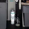 Na zdjęciu znajduje się butelka z napisem "hansgrohe claro swiss IONENTAUSCHER PRO Ion Exchanger Pro" oraz przezroczysta butelka z rozpylaczem, umieszczone w ciemnym wnętrzu szafki. W tle widoczne są elementy instalacji wodnej oraz fragmenty urządzeń kuch