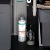 Na zdjęciu znajduje się butelka z napisem "hansgrohe claro HARMONY" oraz przezroczysta butelka z rozpylaczem, umieszczone w ciemnym wnętrzu szafki. W tle widoczne są elementy instalacji wodnej oraz inne przedmioty domowego użytku.