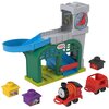Zestaw zabawkowy przedstawia kolorową stację kolejową z niebieskim torem zjazdowym, zieloną bramą i szarą wieżą z zegarem, na której widnieje logo "THOMAS & FRIENDS". Wokół stacji znajdują się różnokolorowe elementy, w tym czerwona lokomotywa z uśmiechnię