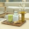Na drewnianej tacy znajduje się szklanka z zielonym napojem, metalowe sitko, bambusowa miotełka, puszka z napisem "PAPER & TEA REVITALIZING MATCHA", pudełko z napisem "MATCHA ABSOLUTE BEGINNER" oraz wazon z suszonymi roślinami. W tle widoczne są jasne meb