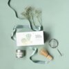 Na zdjęciu znajduje się zestaw do przygotowywania matchy, w skład którego wchodzą: biała prostokątna pudełko z napisem "MATCHA ABSOLUTE BEGINNER", metalowe sitko, bambusowa miotełka oraz puszka z herbatą oznaczona jako "EVERY DAY MATCHA". Obok zestawu leż
