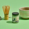 Na zdjęciu znajduje się zestaw do przygotowywania herbaty matcha, w skład którego wchodzą bambusowy pędzel, sitko, puszka z napisem "JAPAN MATCHA" oraz ceramiczna miseczka. Tło jest jednolite, zielone.