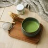 Na zdjęciu znajduje się zestaw do przygotowywania matchy, w skład którego wchodzą miska z zieloną herbatą matcha, bambusowa miotełka, sitko oraz puszka z napisem "WILD & RAW". Wszystkie elementy są ułożone na drewnianej tacy, a w tle widoczny jest szary m