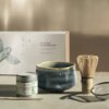 Zestaw do przygotowywania matchy składający się z puszki z napisem "PAPER & TEA EVERYDAY MATCHA", miseczki, bambusowej miotełki, sitka i łyżeczki, umieszczony na tle pudełka z napisem "MATCHA STARTER KIT". Na pudełku widoczna jest ilustracja motyla oraz d