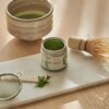 Na zdjęciu znajduje się zestaw do przygotowywania matchy, w skład którego wchodzą miska z zieloną herbatą, otwarty słoik z napisem "PAPER & TEA EVERYDAY MATCHA", bambusowa miotełka oraz sitko i łyżeczka z proszkiem matcha na marmurowej desce. Całość jest 