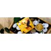 Na zdjęciu znajduje się pudełko kapsułek "Morning Bird Cafe Crema 10 szt." z kolorową grafiką ptaka, umieszczone na tle drewnianej skrzynki wypełnionej białymi kapsułkami oraz filiżanką kawy. Obok pudełka widoczne są zielone liście rośliny.
