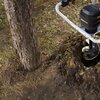Na zdjęciu widać wiertnicę glebową w trakcie pracy obok pnia drzewa, z widocznym logo "WORX" na urządzeniu. Wokół wiertła znajduje się rozkopana ziemia, a w tle widoczna jest trawa.