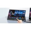 Na zdjęciu widoczny jest monitor ZEUSLAP Z15ST 15" z wyświetlanym kolorowym interfejsem edytora wideo, na którym widać różne klipy i sceny. Na obudowie monitora znajduje się logo "ZEUSLAP", a obok monitora znajduje się ręka wskazująca na ekran.