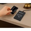 Na zdjęciu widać rękę trzymającą czarną kartę z napisem "EV Charger Card" nad czarnym urządzeniem z logo "Qoltec" i oznaczeniem "RFID" oraz symbolem fali radiowej. Urządzenie jest podłączone kablem, a w tle znajduje się dzwonek hotelowy.