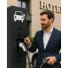 Mężczyzna w garniturze trzyma kartę z napisem "EV Charger Card" przy stacji ładowania pojazdów elektrycznych, na której widnieje symbol samochodu z wtyczką. W tle widoczny jest budynek z napisem "HOTEL".