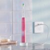 Różowa szczoteczka soniczna PHILIPS Sonicare 4100 HX3689 41 stoi na białej ładowarce na blacie, w tle widoczne są białe płytki ścienne i gniazdko elektryczne. Na szczoteczce znajduje się logo "PHILIPS Sonicare".