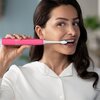 Na zdjęciu widoczna jest różowa szczoteczka soniczna PHILIPS Sonicare 4100 HX3689 41, trzymana przy ustach osoby w białej koszuli. Szczoteczka ma biały, podłużny końcówkę z niebieskimi włoskami.