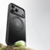 Czarne etui na telefon z trzema widocznymi obiektywami aparatu i okrągłym wycięciem na logo Apple, umieszczone na tle jasnego nieba i fragmentu gałęzi z kroplą wody.