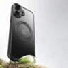Czarne etui TECH-PROTECT Quantum Standlock MagSafe do Apple iPhone 17 jest przedstawione na tle jasnego, rozmytego tła, z widocznym logo Apple na środku. Etui stoi na gałęzi pokrytej mchem, a jego powierzchnia jest gładka i błyszcząca.