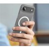 Przezroczyste etui na telefon z widocznym okręgiem MagSafe trzymane w dłoni, z widocznym logo w kształcie jabłka na tylnej części urządzenia. Tło jest rozmyte, w odcieniach szarości i beżu.