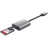 Na zdjęciu znajduje się czytnik kart z logo "Trust" na obudowie, podłączony do kabla USB. Obok czytnika włożona jest karta pamięci z oznaczeniem "64 GB".
