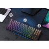 Na zdjęciu znajduje się klawiatura z podświetleniem RGB, umieszczona na ciemnym biurku, obok laptopa i monitora. Wokół klawiatury widoczne są ikony symbolizujące połączenia: Wi-Fi, USB i Bluetooth.